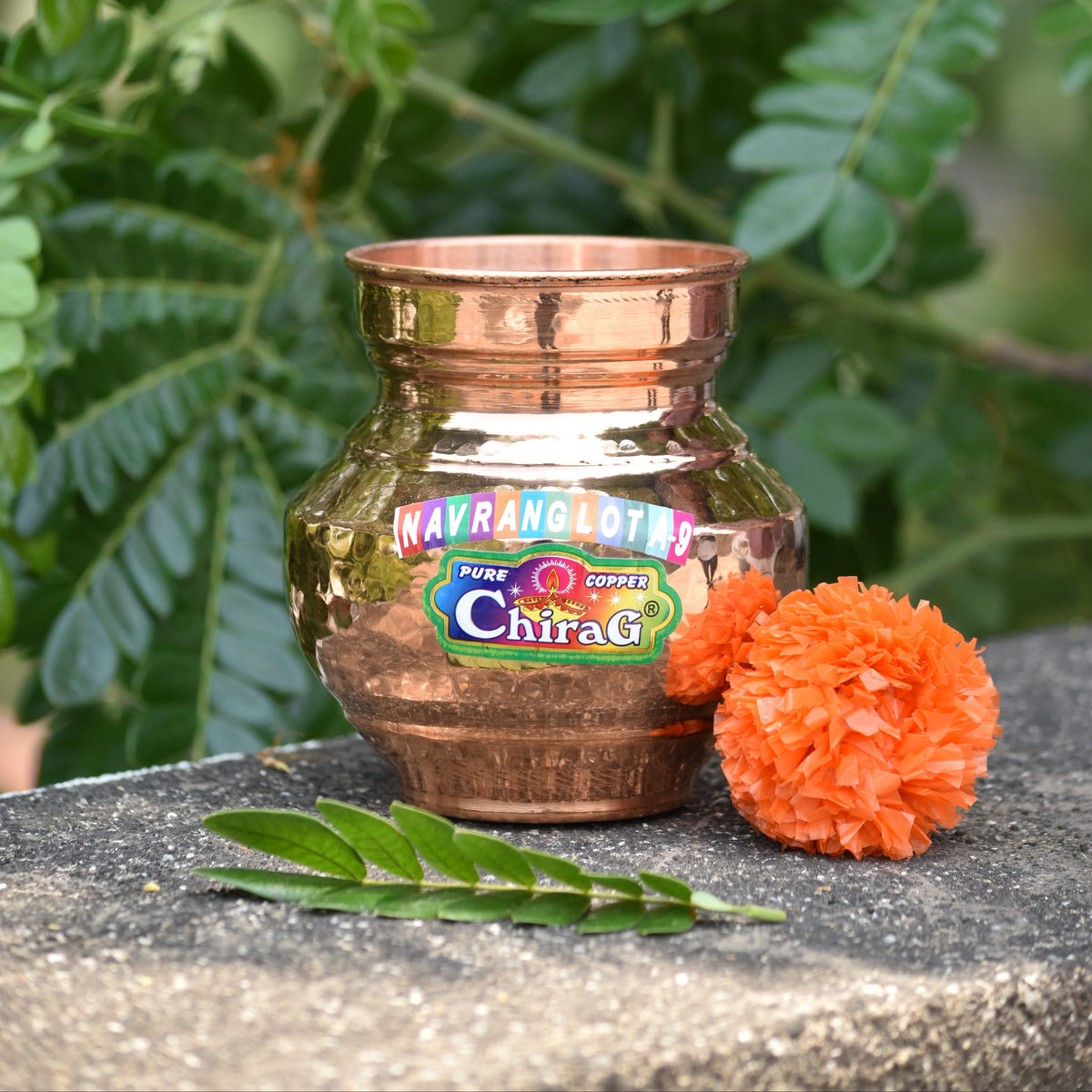 Copper Navrang Lota with a Chirag label on a stone surface with green leaves in the background