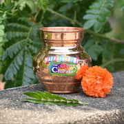 Copper Navrang Lota with a Chirag label on a stone surface with green leaves in the background