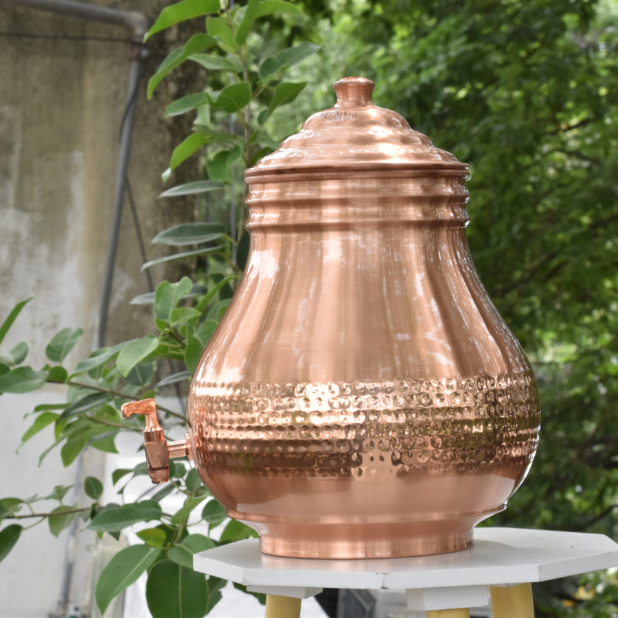 Copper water pot on a stand with greenery in the background
